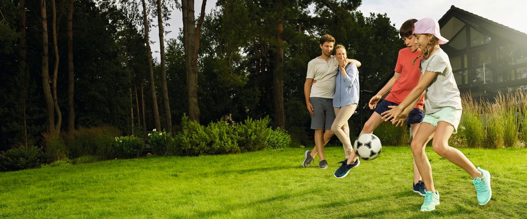 Privathaf tpflichtversicherung: Eine vierköpfige Familie im Garten Tochter und Sohn spielen Fußball. Vater und Mutter schauen im Hintergrund zu.
