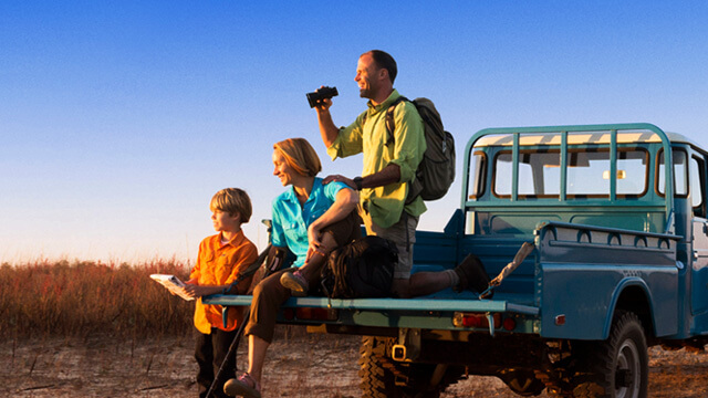 Reisekrankenversicherung. Eine Familie auf Safari: sie sitzen auf einem Pickup und beobachten Tiere.