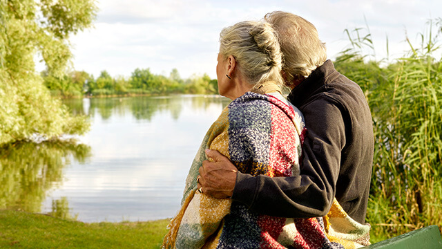 Sterbe­geld­ver­si­che­rung: Ein altes Ehepaar, das bei der ÖVB versichert ist, stehen am Ufer eines Sees und genießen die Natur. 
