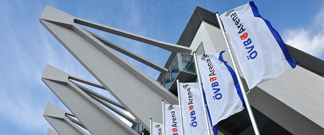 Moderne Architektur der ÖVB Arena mit auffälligen Fensterfronten und wehendem Flaggenbanner. Heller, wolkiger Himmel.