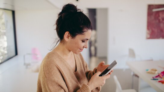 Junge Frau in beiger Strickware schaut konzentriert auf ihr Smartphone in einem modernen, hellen Raum.