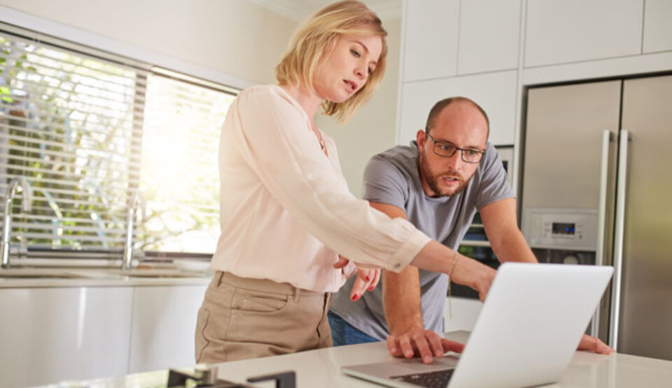 Ein Mann und eine Frau stehen in einer modernen Küche und betrachten gemeinsam den Bildschirm eines Laptops.