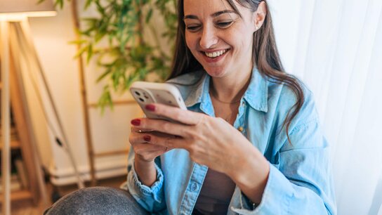 Frau entspannt auf einer Couch und hält ein Smartphone mit einer App-Ansicht in der Hand, lächelnd und zufrieden.