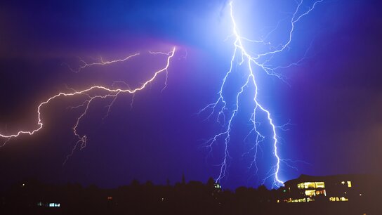 Zwei Blitze erhellen den nächtlichen Himmel über einer Stadt, im Hintergrund sind dunkle Silhouetten von Gebäuden sichtbar.