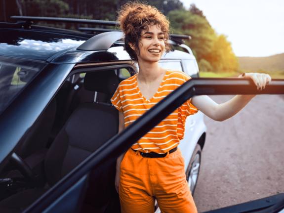 Lächelnde Frau mit lockigem Haar in orange gestreiftem Shirt und orangefarbener Hose steht am offenen Autotür auf einem Landweg.