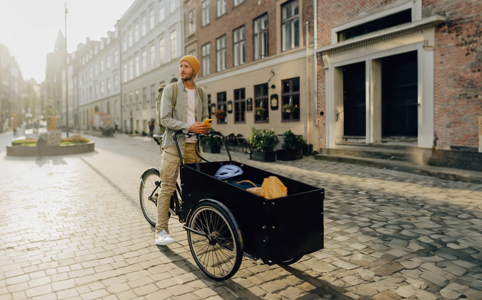 Mann mit gelber Mütze steht neben einem Lastenrad auf einer gepflasterten Straße, umgeben von Gebäuden und Pflanzen.