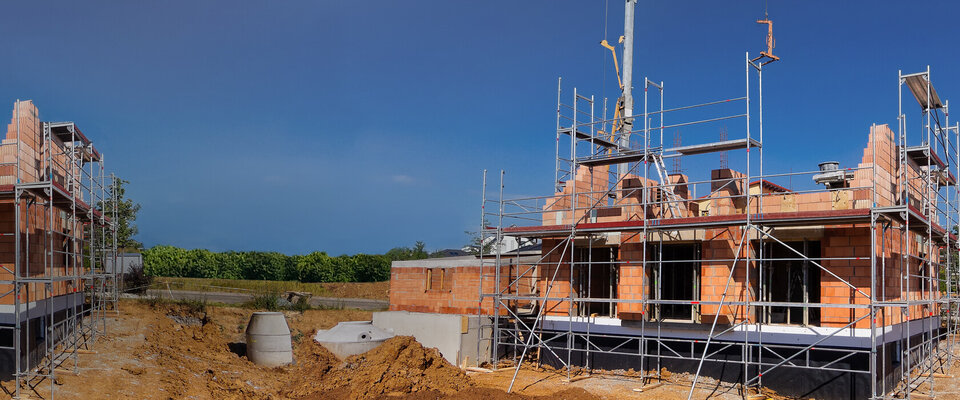 Baustelle mit zwei unvollendeten Häusern, umgeben von Baugerüst und Erdarbeiten unter blauem Himmel.