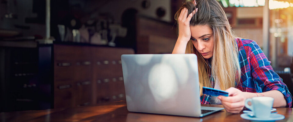 Eine frustrierte junge Frau sitzt an einem Tisch und schaut auf ihren Laptop, während sie eine Kreditkarte in der Hand hält.