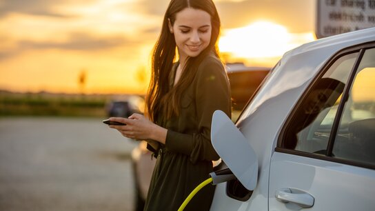 Junge Frau in einem Kleid lädt ein Elektroauto auf und hält ein Smartphone in der Hand, während die Sonne untergeht.