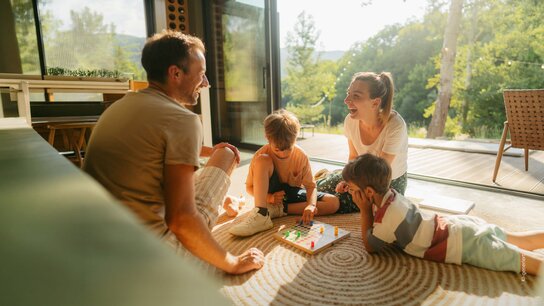 Eine Familie spielt gemeinsam am Boden in einem hellen Raum, umgeben von Naturlicht und Pflanzen.
