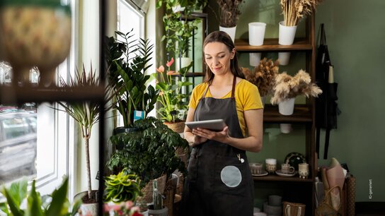 Frau in einem Blumenladen steht mit einem Tablet zwischen Pflanzen und Dekorationen, trägt eine Schürze und lächelt.