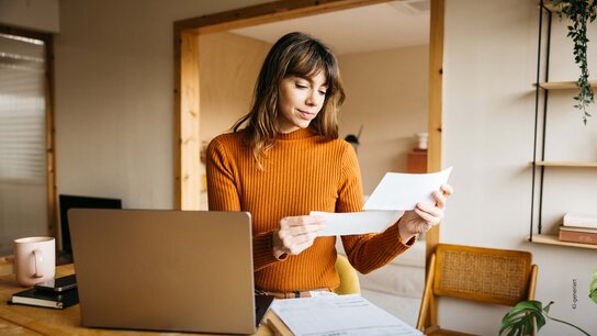 Frau mit einem orangen Pullover sitzt an einem Tisch, schaut sich Dokumente an und arbeitet an einem Laptop.